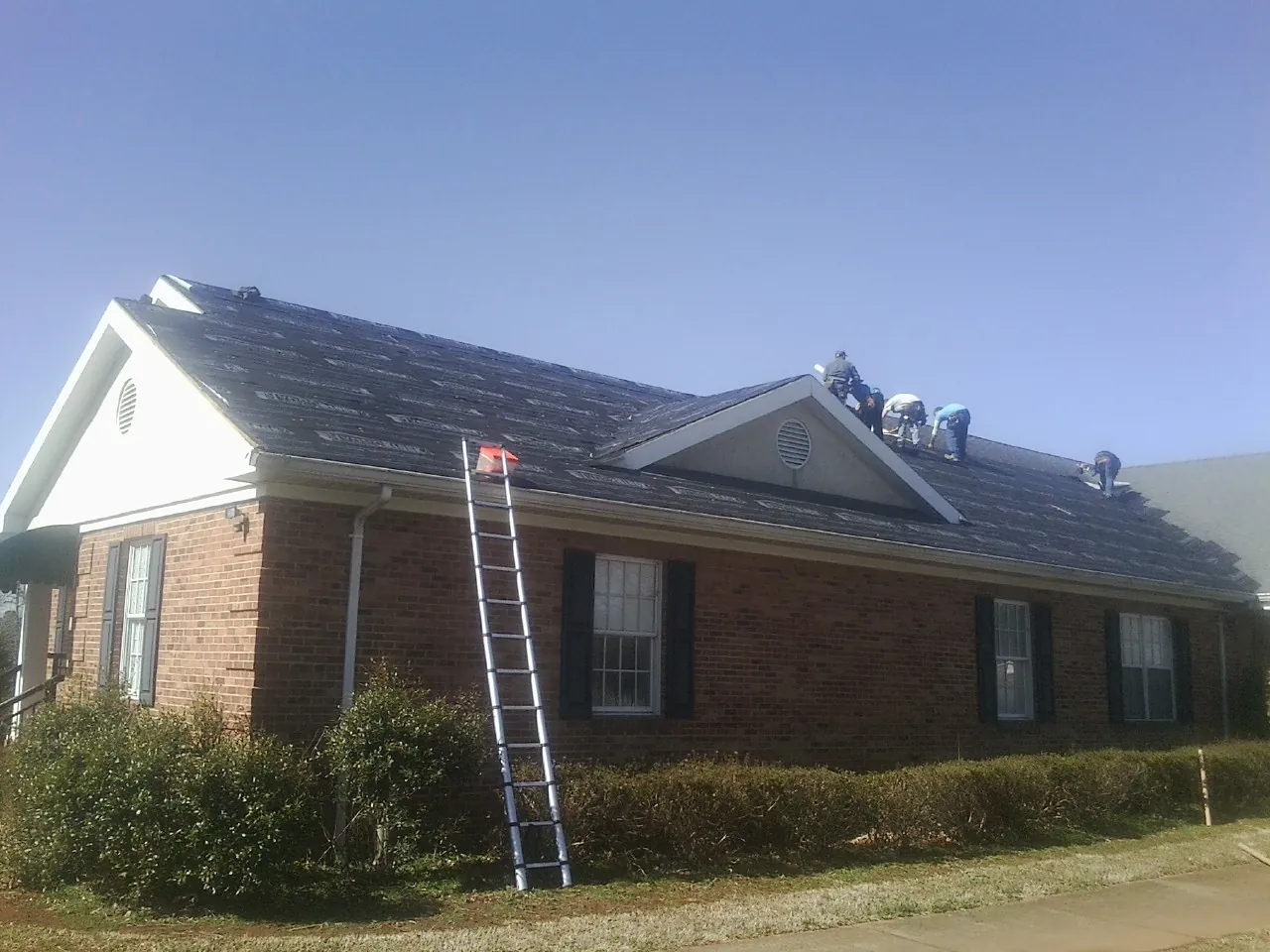 Crew tearing shingles off a brick ranch roof with a ladder set — Metal Roof Repair work by our crew in Reinbeck