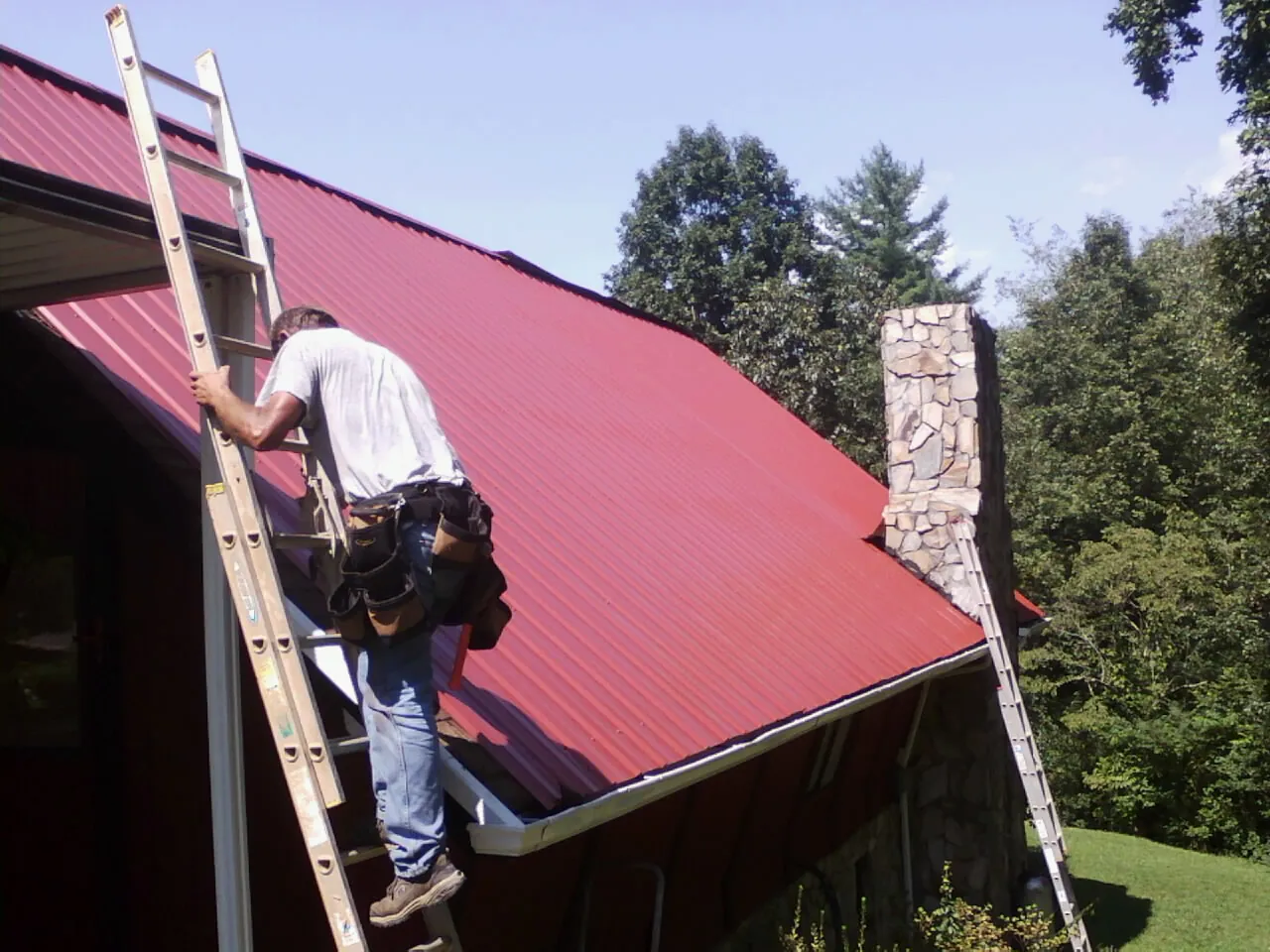 Roofer on a ladder beside a red metal roof and stone chimney — Soffit & Fascia Repair by Roofing Brothers in Reinbeck